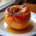 A close-up of a perfectly caramelized baked apples dessert, sitting in a pool of syrup on a white plate.