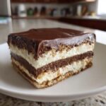 A perfect square slice of chocolate eclair cake showing layers of vanilla pudding, chocolate filling, and graham crackers, topped with chocolate glaze.