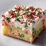 Close-up of a single slice of moist christmas sugar cookie bars topped with thick white frosting and red and green sprinkles.