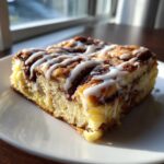 A close-up of a moist slice of cinnamon roll french toast topped with white icing drizzle, sitting on a white plate.