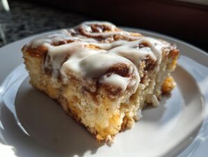 A close-up, sunlit slice of cinnamon roll french toast topped generously with thick white icing.