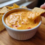 A hand dips a tortilla chip into a small white bowl filled with creamy, orange cottage cheese queso dip with visible red pepper flakes.