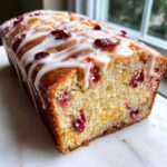 A close-up of a freshly baked cranberry orange cake loaf drizzled with white glaze and studded with dried cranberries.