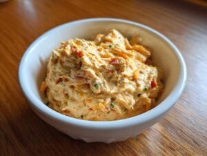 A close-up of the creamy, orange-colored boat dip mixed with shredded cheese and red peppers in a white serving bowl.