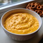 Close-up of a thick, orange beer cheese dip served in a white bowl, with pretzels visible in the background.