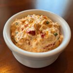 Close-up of creamy, orange-hued boat dip mixed with red peppers and green chilis in a small white bowl.