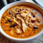 Close-up of a bowl of rich, orange-hued creamy chicken enchilada soup filled with shredded chicken, black beans, and corn.