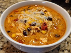 Close-up of a white bowl filled with rich, orange creamy chicken enchilada soup topped with shredded chicken, black beans, corn, and melted cheese.