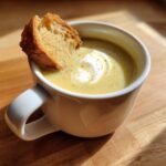 A close-up of creamy garlic soup in a white mug, topped with a swirl of cream and a toasted crouton.