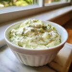 A close-up of creamy, chunky pickle dip mixed with dill, served in a white bowl near a window.