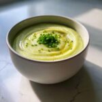 A close-up of a bowl of creamy, pale green potato leek soup topped with fresh chives and olive oil.