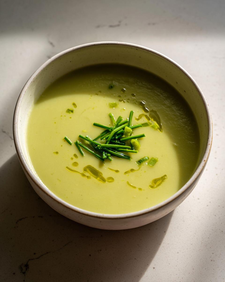 A close-up of a bowl of smooth, pale green potato leek soup garnished with fresh chives and olive oil.