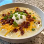 A close-up of a creamy bowl of potato soup recipe topped with sour cream, bacon bits, shredded cheddar cheese, and green onions.