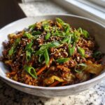 A close-up of a savory egg roll in a bowl featuring ground meat, cabbage, and carrots, garnished with fresh green onions and sesame seeds.