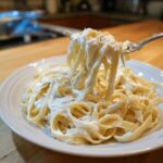 A fork lifting fettuccine noodles generously coated in creamy olive garden alfredo sauce from a white plate.