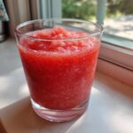 A close-up of a vibrant, frozen strawberry daiquiri served in a clear glass, sitting on a sunlit windowsill.