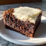 Close-up of a rich, dark square of gingerbread brownies topped with thick white cream cheese frosting.