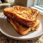 Two thick slices of golden brown sourdough french toast stacked on a light plate, catching bright sunlight.