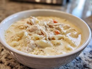 A close-up of a white bowl filled with rich homemade cream of chicken soup, featuring shredded chicken, egg noodles, and carrots.