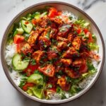 Overhead view of a vibrant bowl featuring saucy harissa chicken served over white rice with fresh lettuce, cucumbers, and tomatoes.