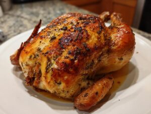 A juicy, golden-brown whole chicken roasted with herbs resting on a white platter.