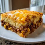 A square serving of Mexican cornbread casserole featuring a layer of seasoned ground beef and corn topped with melted cheddar cheese.