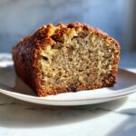 Close-up of a thick slice of moist banana bread recipe showing the crumb texture and walnuts, served on a white plate.