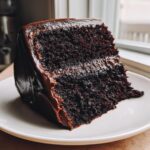 A thick, moist slice of dark chocolate fudge cake covered in rich, glossy chocolate frosting, sitting on a white plate.
