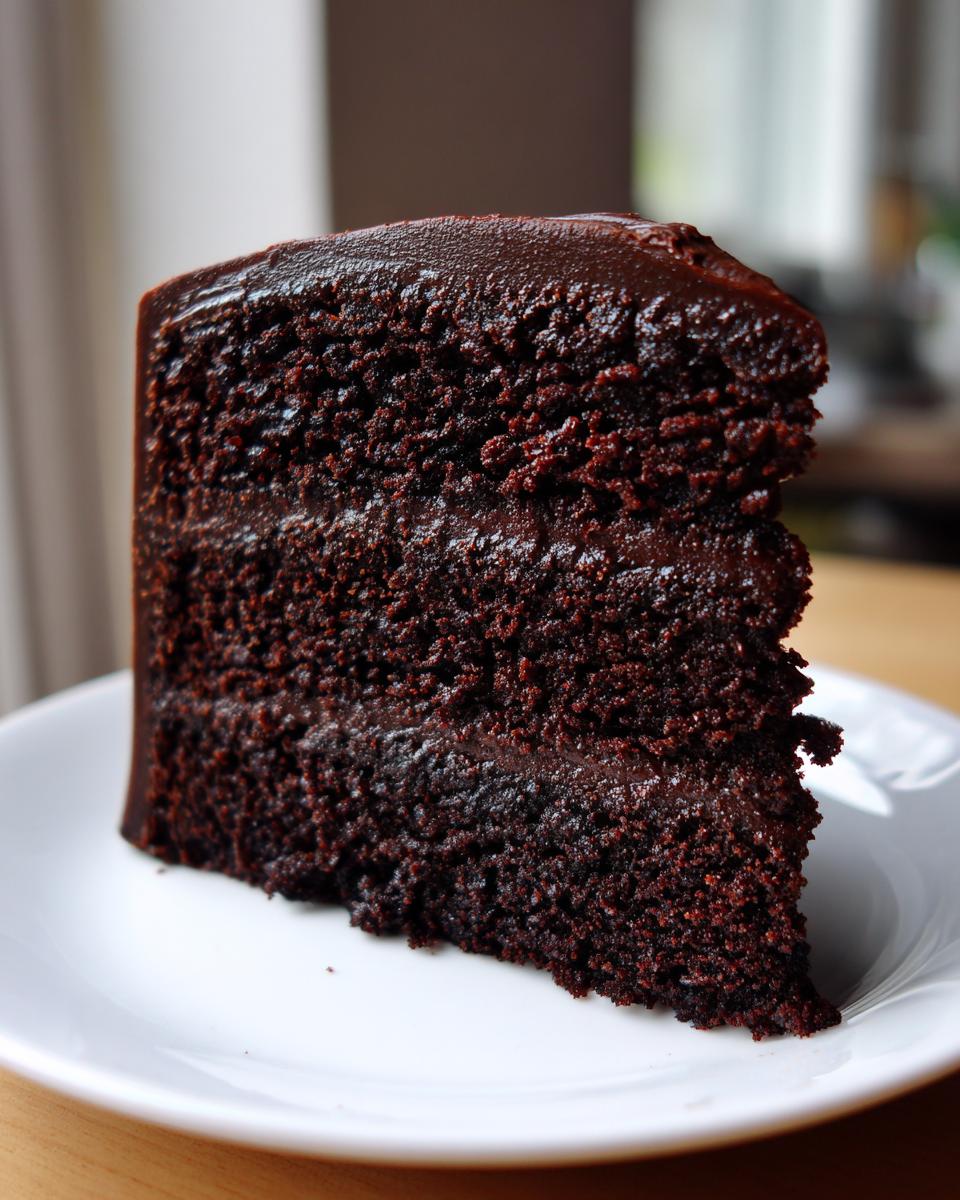 A tall, rich slice of moist chocolate fudge cake with dark ganache frosting on a white plate.