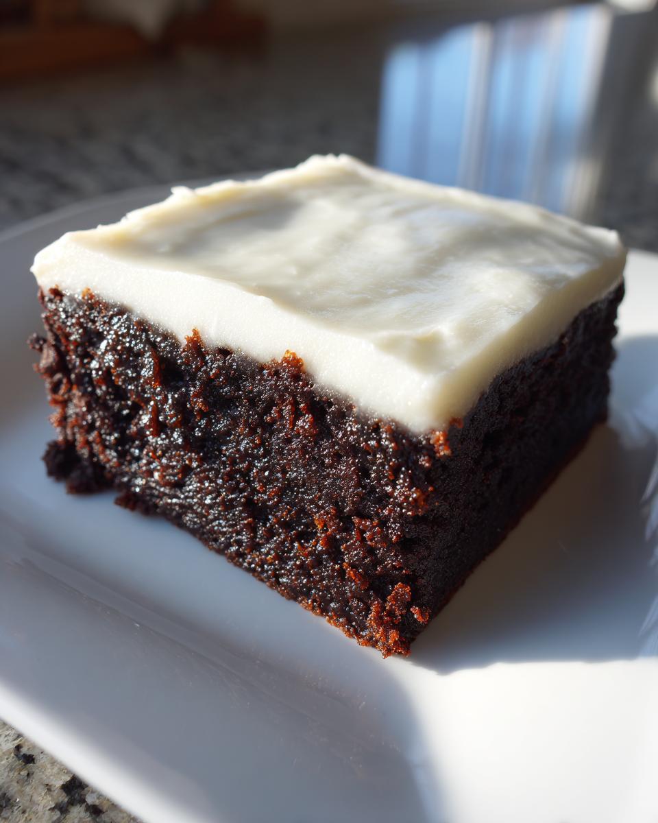 A close-up shot of a square slice of rich, dark gingerbread brownies topped with thick white frosting.