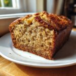 A close-up of a moist slice of greek yogurt banana bread with a caramelized, sugary crust, sitting on a white plate.