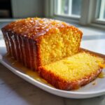 A moist orange juice cake loaf, partially sliced, covered in a thick, dripping orange glaze.