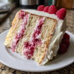 A tall slice of moist white chocolate raspberry cake showing layers of white cake, raspberry filling, and white frosting, topped with fresh raspberries.
