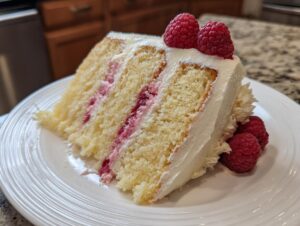 A moist slice of white chocolate raspberry cake with layers of yellow cake, raspberry filling, and white frosting, topped with fresh raspberries.