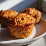 Three freshly baked morning glory muffins with visible shredded carrots, sitting on a light-colored plate in bright sunlight.