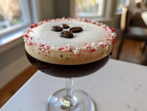 Close-up of a peppermint espresso martini with a thick foam top, crushed candy cane rim, and three coffee beans.