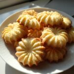 A close-up of several golden, piped butter cookie treats stacked on a white plate, bathed in sunlight.