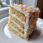 A tall, decadent slice of homemade coconut cake recipe with four layers of cake and frosting, topped with toasted coconut flakes.