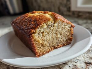 Close-up of a thick slice of homemade banana bread recipe with a dark golden crust, sitting on a white plate.