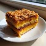 A close-up of a square slice of three-layer pumpkin crunch dessert with a crumbly topping on a white plate.