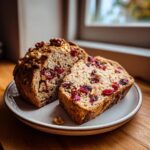 A loaf of cranberry walnut bread, partially sliced, showing bright red cranberries and walnuts throughout the crumb.