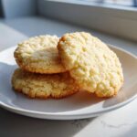 A stack of three golden, crumbly soft sugar cookies dusted with sugar, resting on a white plate.