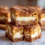 Three rich, square bars of sopapilla cheesecake stacked on a marble surface, showing a creamy white filling and cinnamon sugar topping.