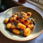 A bowl of pan-seared sweet potato gnocchi topped with crispy sage leaves and grated cheese.