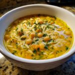 Close-up of a bright yellow chickpea soup featuring chickpeas, orzo pasta, carrots, and fresh parsley.