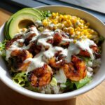 Close-up of a vibrant shrimp bowl featuring grilled shrimp, corn, avocado, and a creamy white sauce over rice and lettuce.