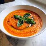 A close-up of a bowl of vibrant orange tomato basil soup, garnished with fresh basil leaves and a drizzle of olive oil.