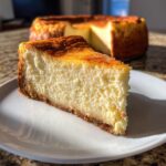 A thick slice of creamy white chocolate cheesecake with a golden-brown top and graham cracker crust, served on a white plate.
