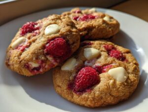 Three freshly baked white chocolate raspberry cookies piled on a white plate, showing melted white chocolate chips and bright red raspberries.
