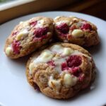 Three freshly baked white chocolate raspberry cookies with melted white chocolate drizzle on a white plate.
